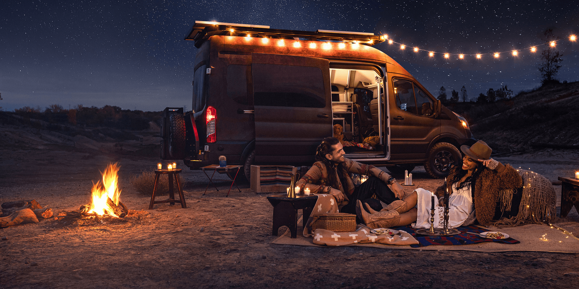 A man an woman sitting by a fire at night next to a Ford Transit campervan. String lights are are lit on the Transit.