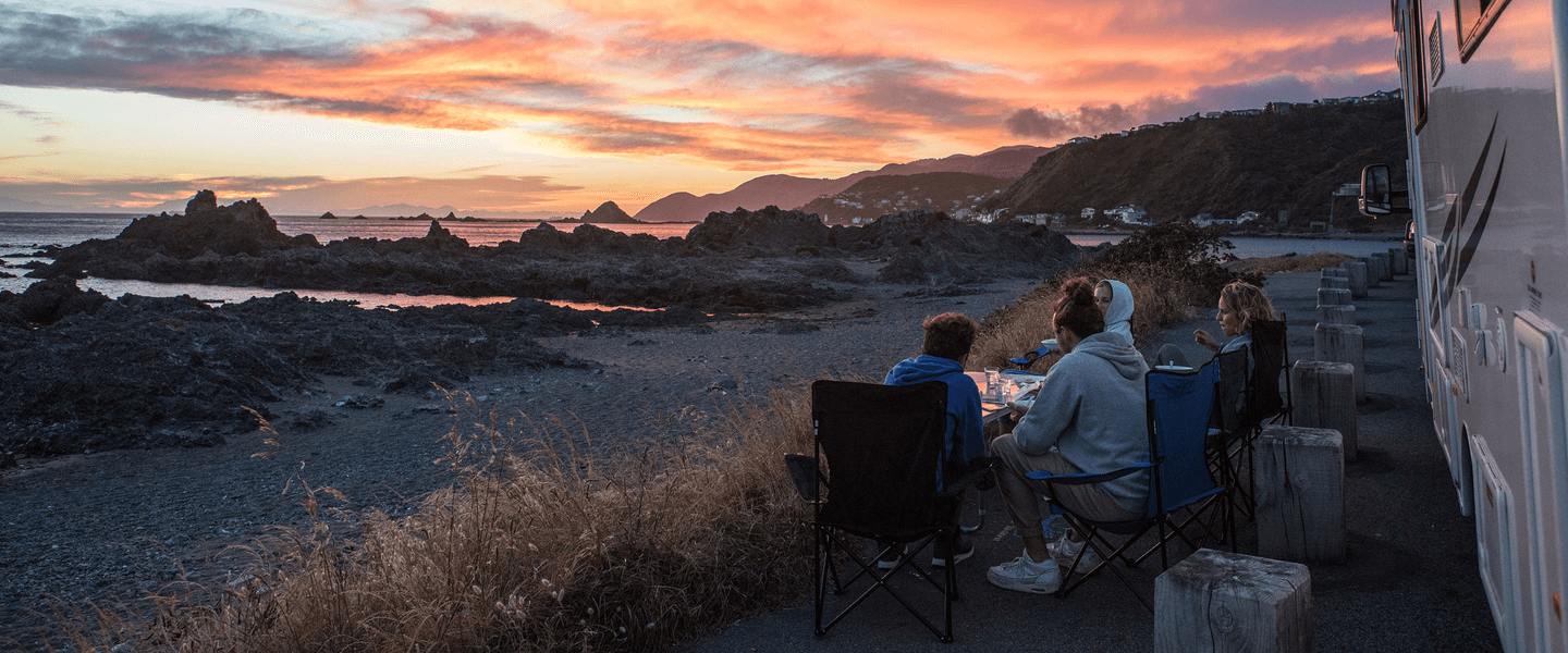 An RV is parked near the coast. Several people are sitting in chairs next to it watching the sunset.