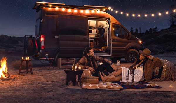 A man an woman sitting by a fire at night next to a Ford Transit campervan. String lights are are lit on the Transit.